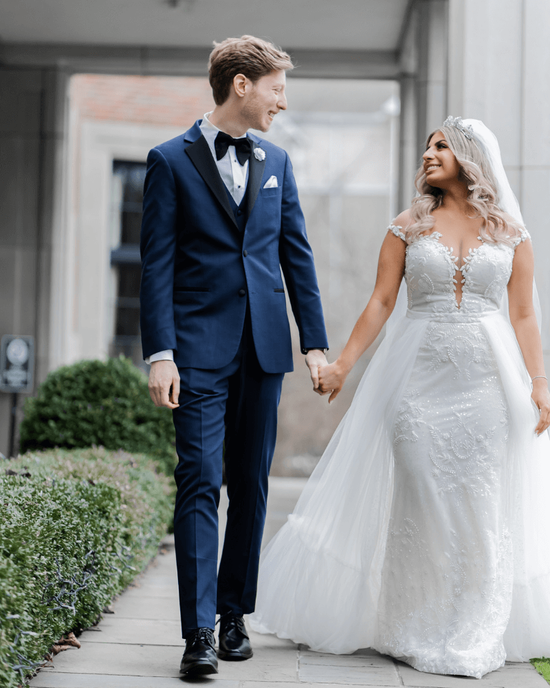 Bride and groom hold hands. Groom in blue suite. Bride in off shoulder gown with overskirt.