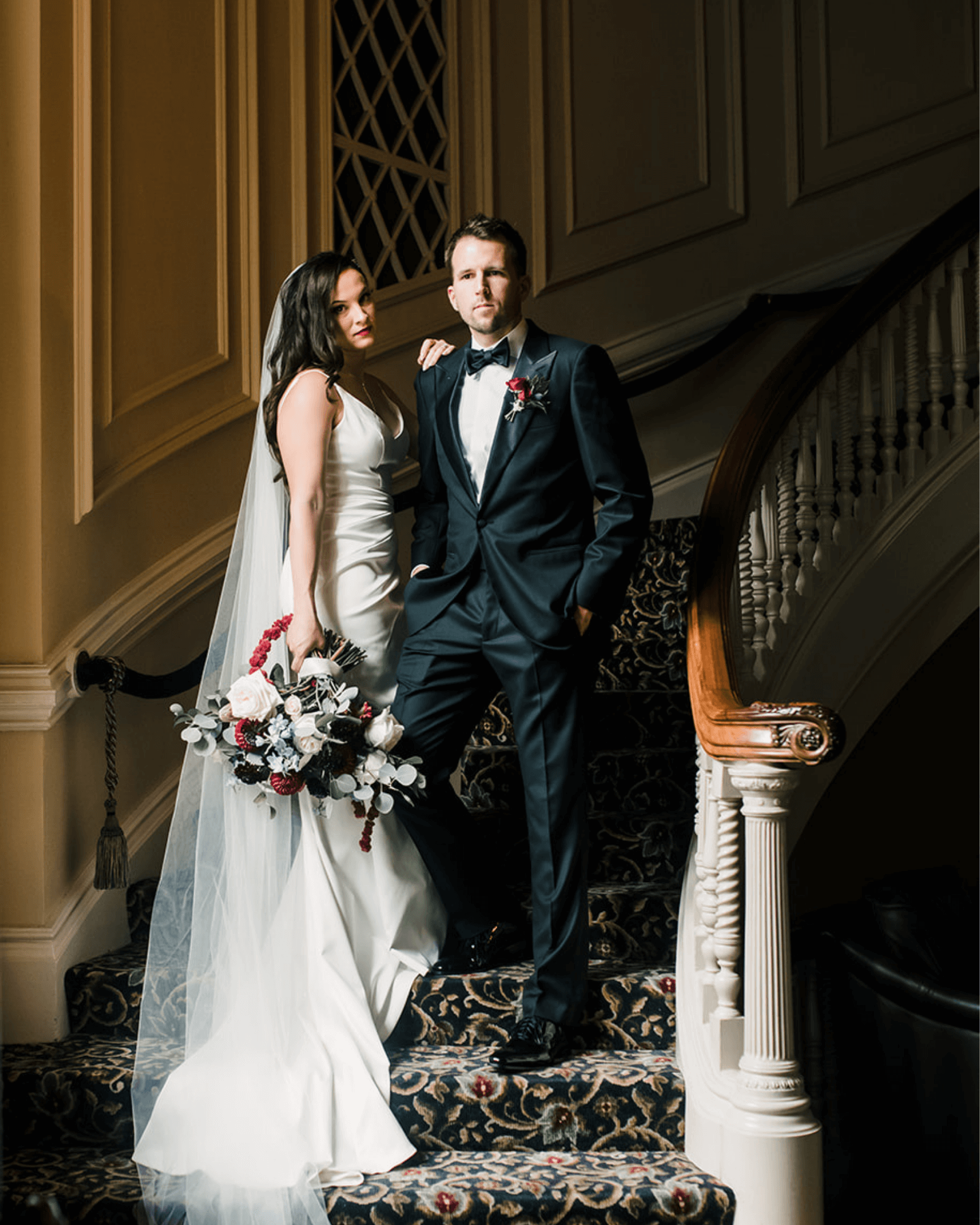 Bride Lauren with Husband on staircase. Husband in dark suite with hands in pockets. Lauren in white gown with veil behind her