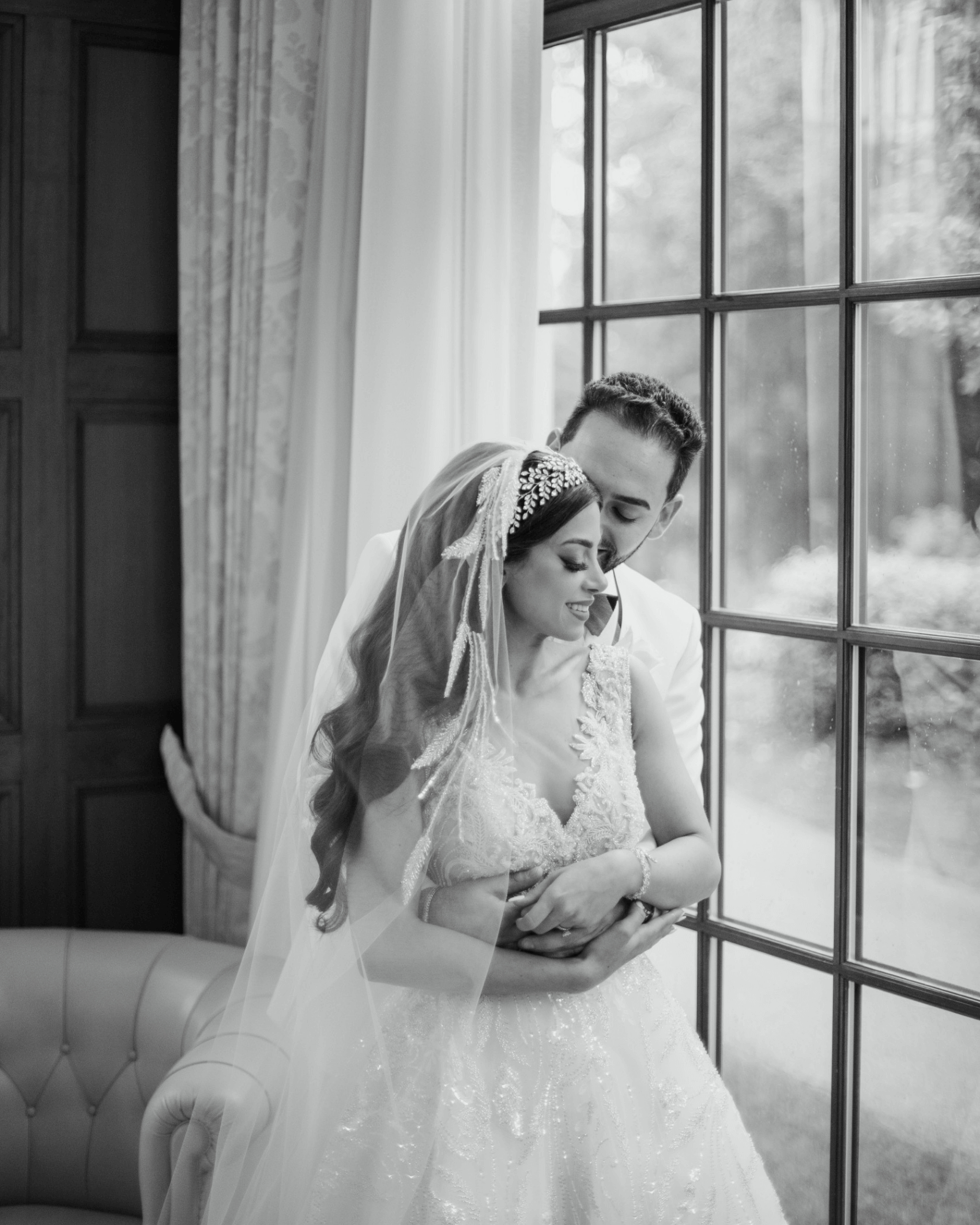 Bride and groom standing by window. Bride in a line gown. Groom hugging bride from behind.