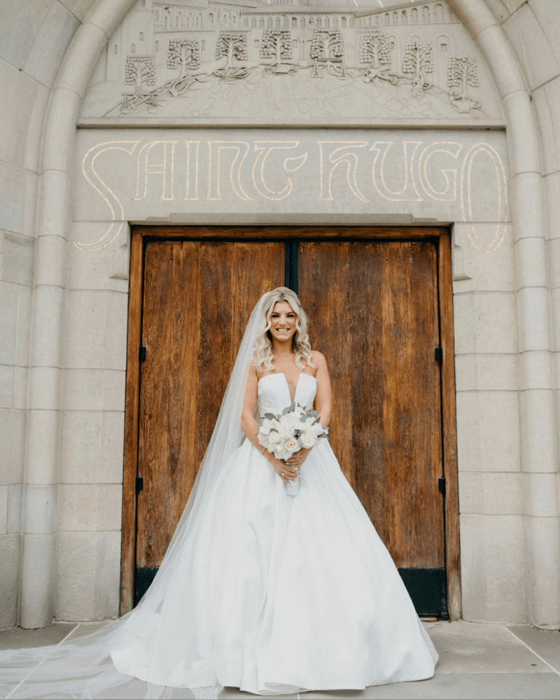 Gina, Bride Standing in front of wooden door. Bridal is wearing a ballgown and holding a bouquet.