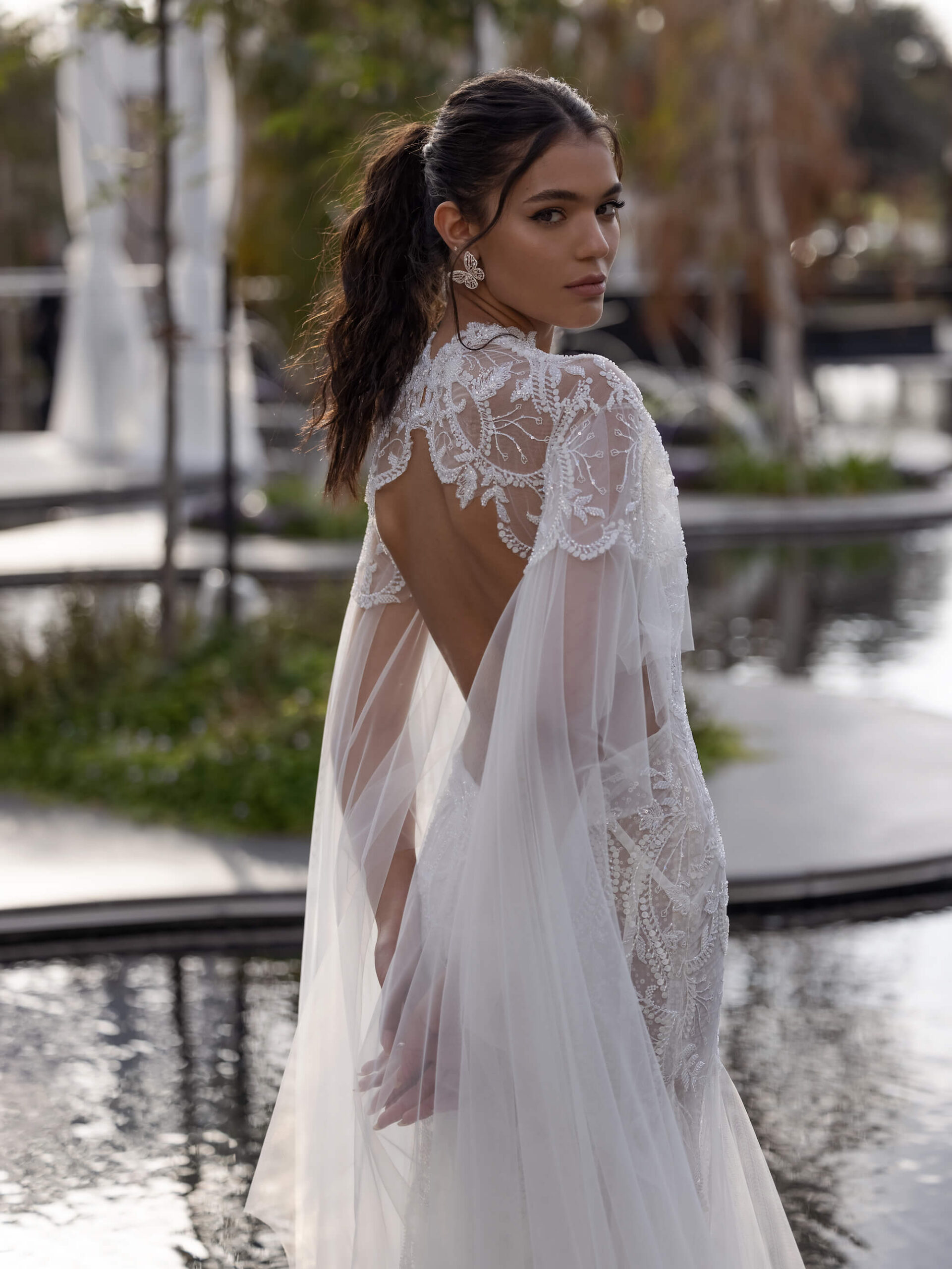 Bride looks over her right shoulder. Adorned appliqué lines the shoulders and opens into a tulle cape. From the back, we see a bond